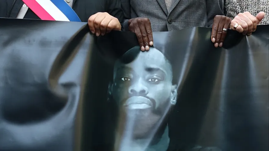 Los asistentes y las autoridades sostienen una pancarta con un retrato mientras participan en una marcha silenciosa en homenaje a Pengo Bossio Wangi, un hombre que murió tras recibir disparos de un atacante el 23 de octubre; la marcha fue organizada por su club de MMA cerca del ayuntamiento de Aubervilliers, al norte de París, el 25 de octubre de 2026.