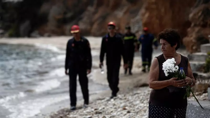 Varios bomberos (detrás) buscan a desaparecidos mientras una mujer sostiene un ramo de flores tras el incendio forestal de Mati, al noreste de Atenas (Grecia), hoy 27 de julio de 2018. El número de víctimas mortales por los graves incendios ocurridos el lunes en Grecia ha ascendido ya a 86, según ha informado el jefe del servicio forense de Atenas, Nikos Karakukis.